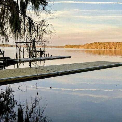 A wooden dock extends over calm lake water, with trees and vegetation at the shoreline and in the distance under a pastel sky with streaks of clouds.