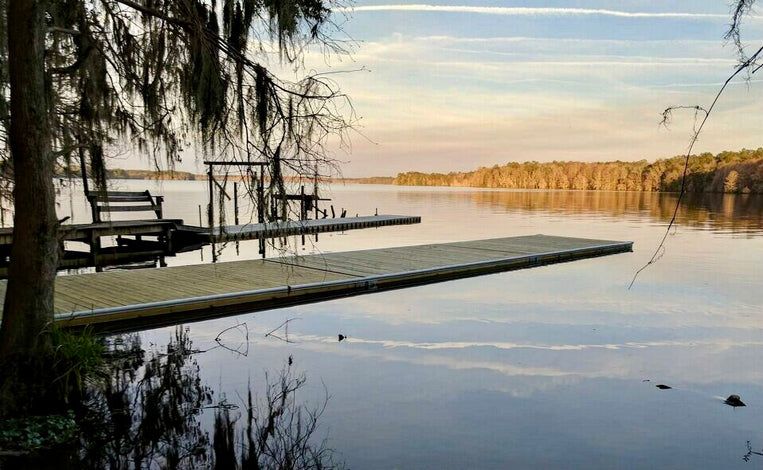 A wooden dock extends over calm lake water, with trees and vegetation at the shoreline and in the distance under a pastel sky with streaks of clouds.